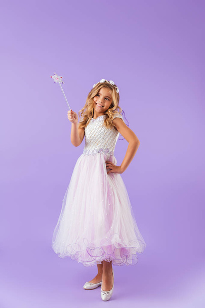 Full length portrait of a smiling pretty girl dressed in a princess dress isolated over violet background, holding magic wand - Fotó, kép