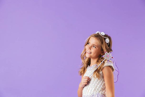 Portrait of a smiling cute pretty girl dressed in a princess dress isolated over violet background, holding magic wand, looking away - Φωτογραφία, εικόνα
