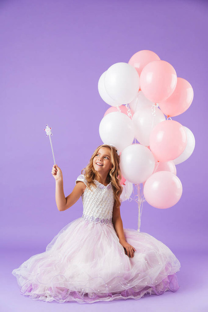 Pretty little girl dressed in princess dress sitting isolated over violet background, holding bunch of balloons and a magical wand - Fotó, kép