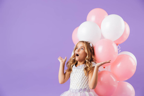 Cute pretty little girl dressed in princess dress isolated over violet background, holding bunch of balloons, looking at copy space - Fotó, kép