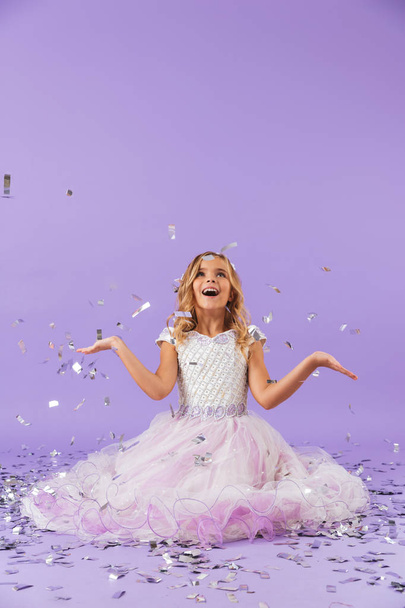 Pretty little girl dressed in princess dress sitting isolated over violet background - Foto, afbeelding