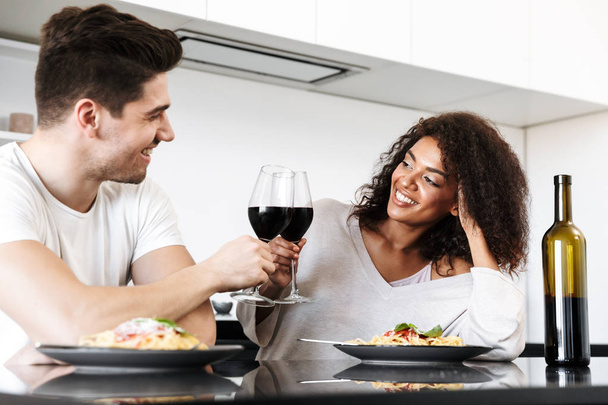 Beautiful young multiethnic couple having a romantic dinner at home, drinking red wine and eating pasta, toasting - 写真・画像