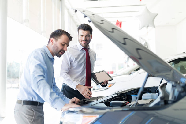 Smiling dealer holding digital tablet while looking at client analyzing car engine - Foto, imagen