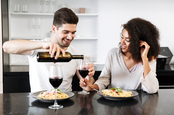 Beautiful young multiethnic couple having a romantic dinner at home, drinking red wine and eating pasta, pouring - Foto, Imagem