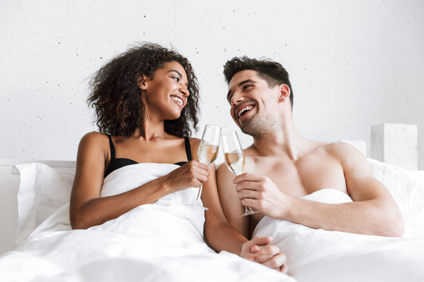 Beautiful happy young multiethnic couple relaxing in bed under blanket, drinking sparkling champagne from glasses - Фото, зображення