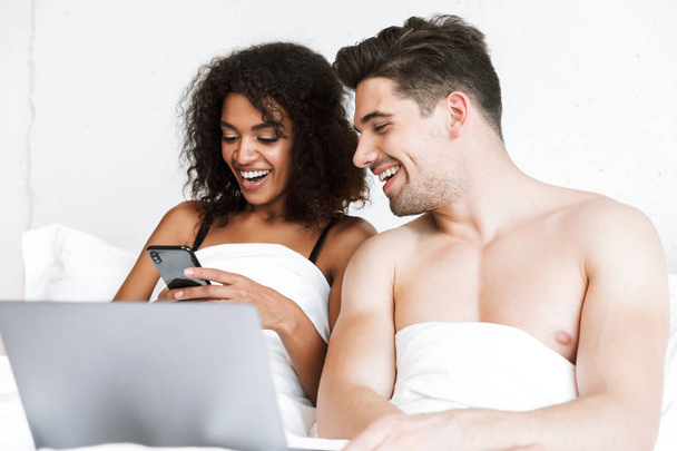 Beautiful happy young multiethnic couple relaxing in bed under blanket, man using laptop computer, woman holding mobile phone - Zdjęcie, obraz