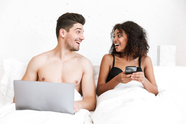 Beautiful happy young multiethnic couple relaxing in bed under blanket, man using laptop computer, woman holding mobile phone - Valokuva, kuva