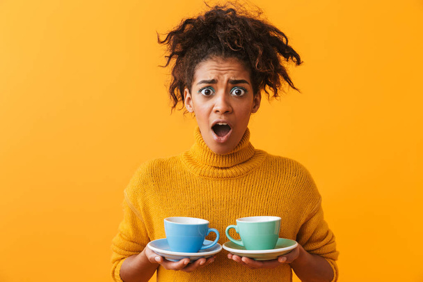 Confused african woman wearing sweater holding cups on saucers isolated over yellow background - Φωτογραφία, εικόνα