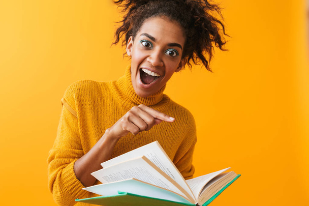 Cheerful african woman wearing sweater holding a book isolated over yellow background - Foto, afbeelding