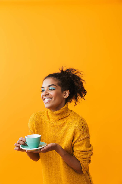 Cheerful african woman wearing sweater holding cup on a saucer isolated over yellow background - Foto, imagen
