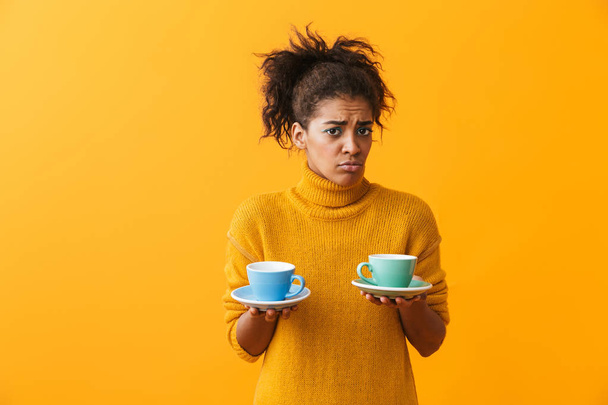 Confused african woman wearing sweater holding cups on saucers isolated over yellow background - Foto, afbeelding