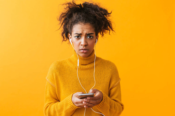 Upset african woman wearing sweater standing isolated over yellow background, listening to music with earphones, holding mobile phone - Foto, imagen