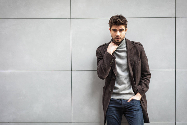 young attractive man in coat posing on a grey wall , stylish man - Foto, afbeelding