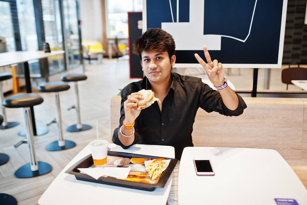 Stylish indian man sitting at fast food cafe and eating hamburger and gesture peace sign hand. - Fotoğraf, Görsel