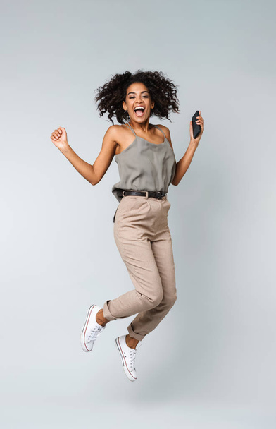 Full length of a happy young african woman casually dressed jumping isolated over gray background, holding mobile phone - Foto, afbeelding