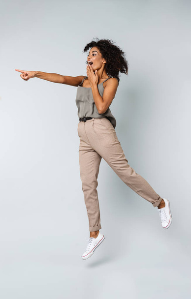 Full length of a happy young african woman casually dressed jumping isolated over gray background, pointing at copy space - Fotó, kép