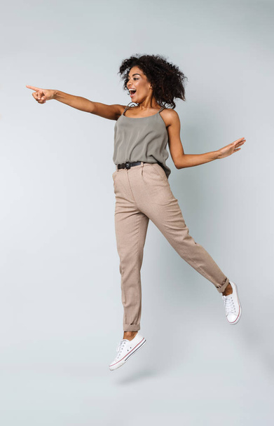 Full length of a happy young african woman casually dressed jumping isolated over gray background, pointing at copy space - Фото, изображение