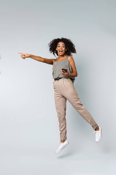 Full length of a happy young african woman casually dressed jumping isolated over gray background, pointing at copy space - Foto, immagini