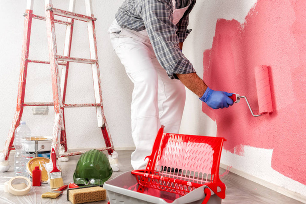 Кавказский маляр в белом рабочем комбинезоне, с роллером, раскрашивающим стену красочной живописью. Строительная промышленность
. - Фото, изображение