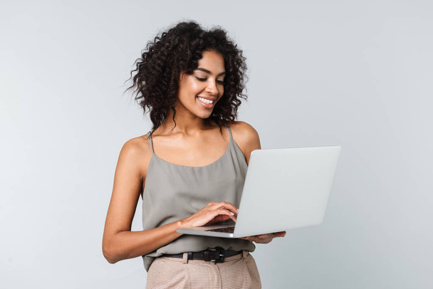 Full length of a happy young african woman casually dressed standing isolated over gray background, working on laptop computer - Photo, image