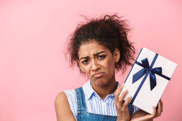 Beautiful disappointed young african woman holding gift box over pink background - Φωτογραφία, εικόνα