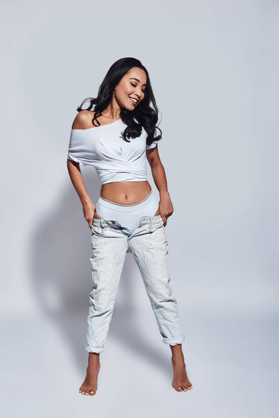 Full length of beautiful young woman smiling and adjusting her jeans while standing against grey background - Foto, immagini