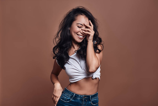 Shy beauty, Beautiful young woman smiling and keeping eyes closed while standing against brown background - Фото, изображение