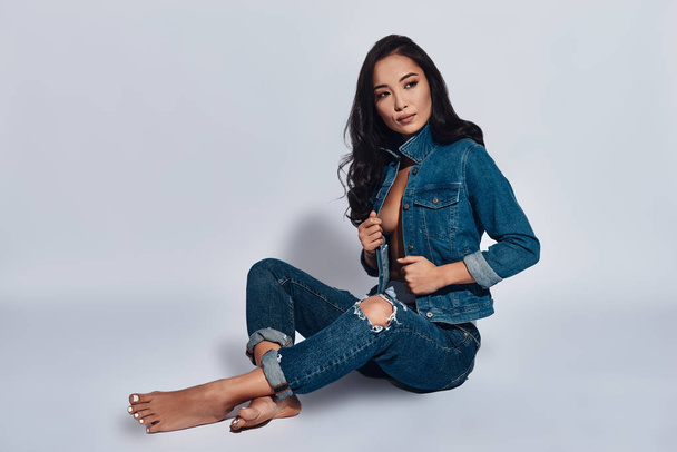 mixed race brunette woman wearing jeans and posing in studio, sitting on floor and looking at camera  - 写真・画像