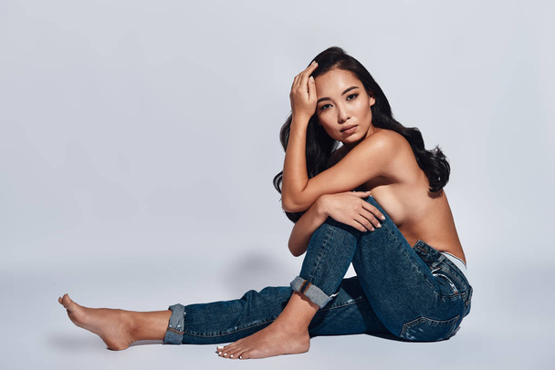 shirtless brunette woman wearing jeans and posing in studio, sitting on floor and looking at camera  - Photo, image