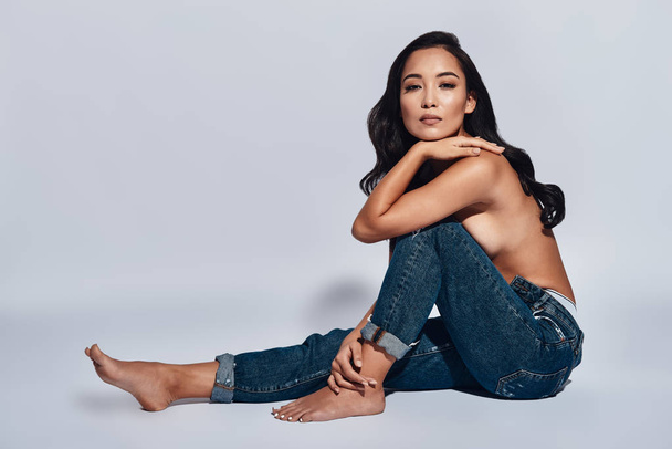 shirtless brunette woman wearing jeans and posing in studio, sitting on floor and looking at camera  - Foto, Imagem