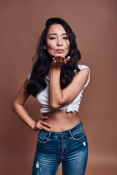 Kiss for you, Beautiful young woman blowing a kiss and smiling while standing against brown background - Fotoğraf, Görsel