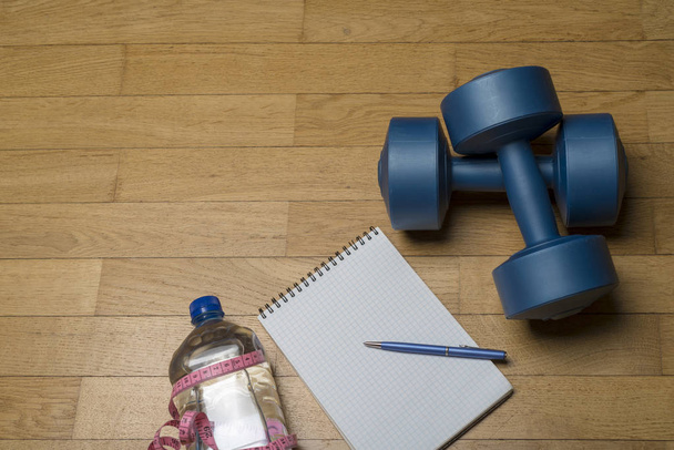 Entraînement, exercice, gaieté et santé - deux haltères en plastique, un cahier, de l'eau minérale et un stylo sur le sol en bois. Le concept d'un mode de vie sain, fitness
. - Photo, image