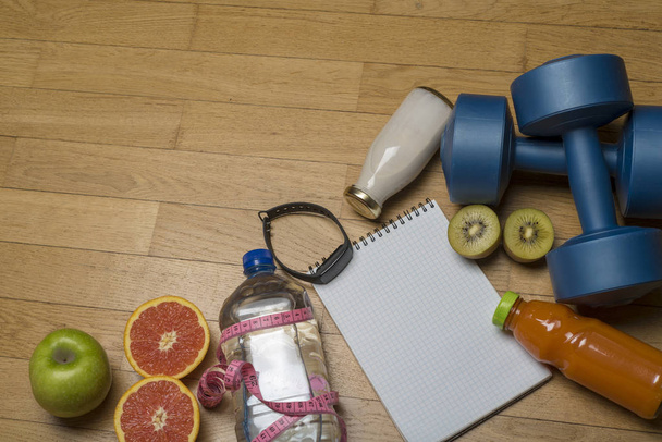 Training, exercise, cheerfulness and health - two plastic dumbbells, a notebook, mineral water with juice, fruit and a pen on the wooden floor. The concept of a healthy lifestyle, fitness. - Photo, Image