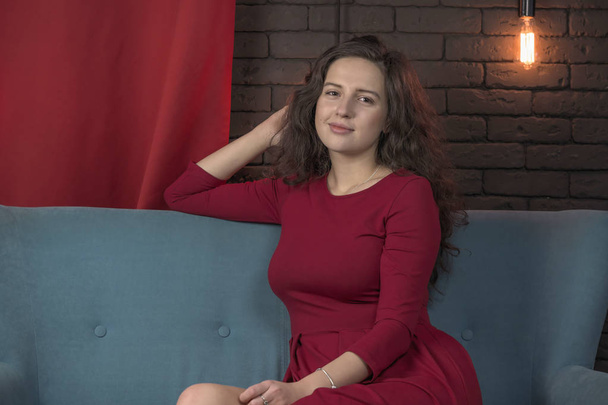 Beautiful attractive girl in a red dress sitting on a blue sofa - Fotó, kép