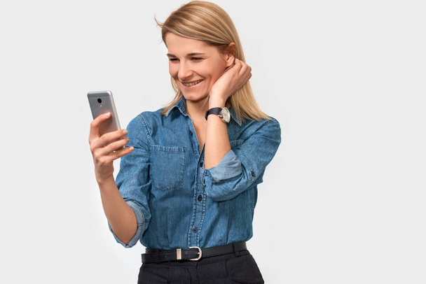 Portrait of cheerful blonde woman, holds modern smart phone, feeling happy to recieve good news, dressed in denim shirt, poses against white studio background with blank space for your advertisement  - Photo, Image