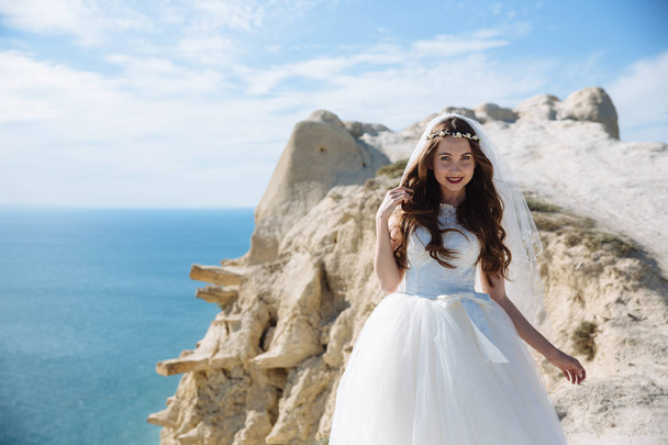 Beautiful happy bride in wedding dress and veil posing on rock background of ocean - Photo, image