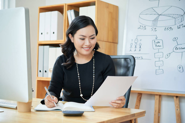 Sonriente mujer empresaria asiática leyendo documento y tomando notas en planificador
 - Foto, imagen