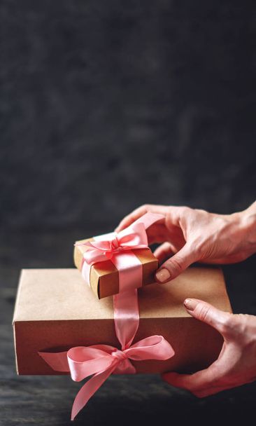 Руки, що тримає подарункові коробки упаковані в Крафт-папір з рожевою стрічкою на темному тлі дерев'яні. Концепція святкова листівка для закоханих і дню жіночий день. Місце для тексту - Фото, зображення
