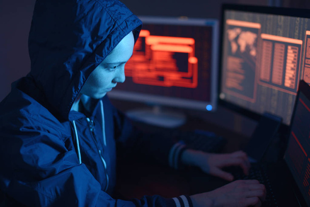 Girl hacker in a hood typing program code while committing a cybercrime hacking a system firewall on the background of screens in the dark under neon light. Cyber security concept - Foto, Imagem