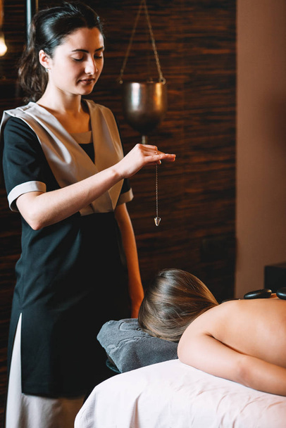 Specialist making biolocation with crystal pendulum. Young charming girl on a panchakarma procedure laying on a massage table. beautiful woman spending time at modern spa cabinet relaxing. Soft yellow light. - Foto, imagen