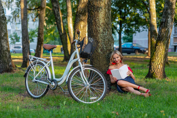 A young blonde woman with a white bike is sitting in a park near a tree and reading a book - image - Fotoğraf, Görsel