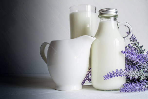 Vintage glass bottle with milk, next to a beautiful jug and glass of milk and a branch of lavender flowers on a white vintage background - Fotoğraf, Görsel