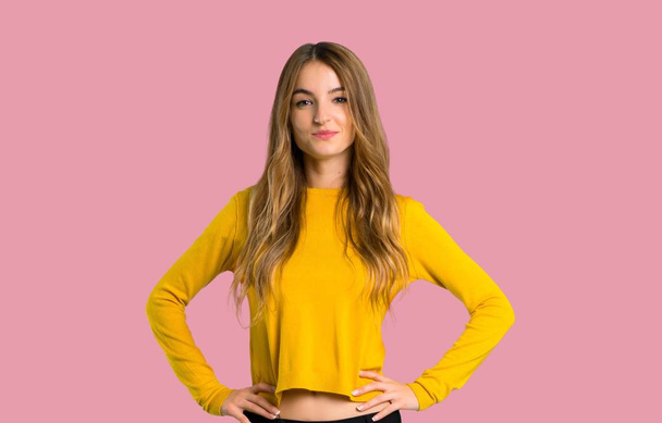 niña con suéter amarillo posando con los brazos en la cadera y riendo mirando hacia el frente sobre un fondo rosa aislado
 - Foto, imagen