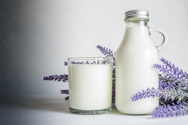 Vintage glass bottle with milk, glass of milk and a branch of lavender flowers on a white vintage background - 写真・画像