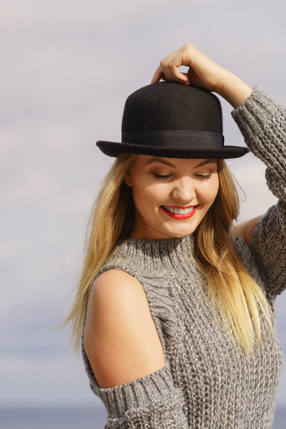 Beautiful young blonde woman wearing fedora hat and sweater jumper with holes on shoulders posing outdoor - 写真・画像