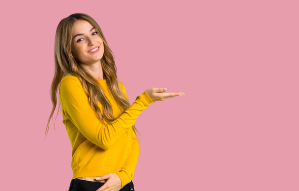 junges Mädchen mit gelbem Pullover, das eine Idee präsentiert, während es lächelnd in Richtung auf isoliertem rosa Hintergrund blickt - Foto, Bild