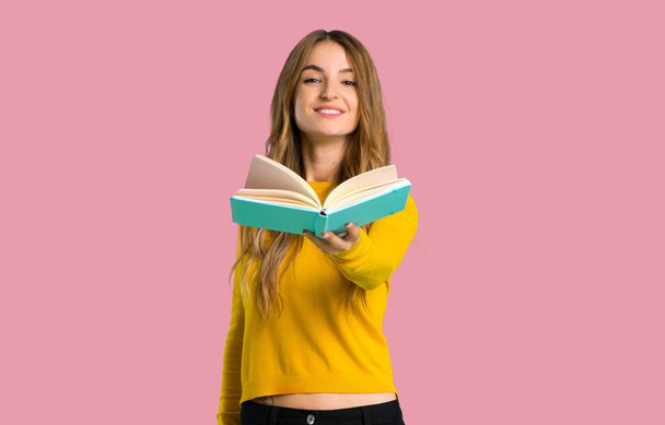 young girl with yellow sweater holding a book and giving it to someone on isolated pink background - 写真・画像