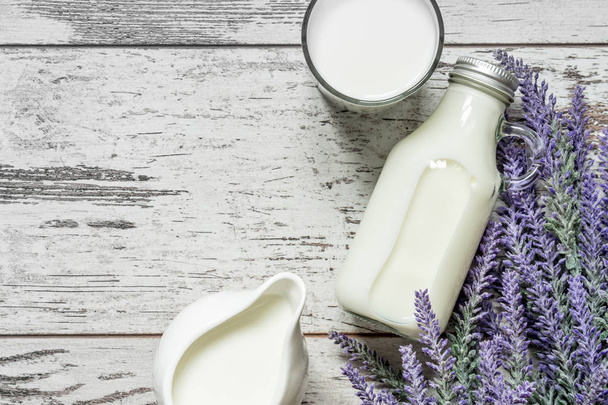 Vintage glass bottle with milk, a glass with milk and a beautiful white jug next to a branch of lavender flowers on a vintage wooden background. View from above. - Photo, Image