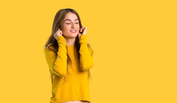 молодая девушка в жёлтом свитере закрывает уши руками. Разочарованное выражение на изолированном желтом фоне
 - Фото, изображение
