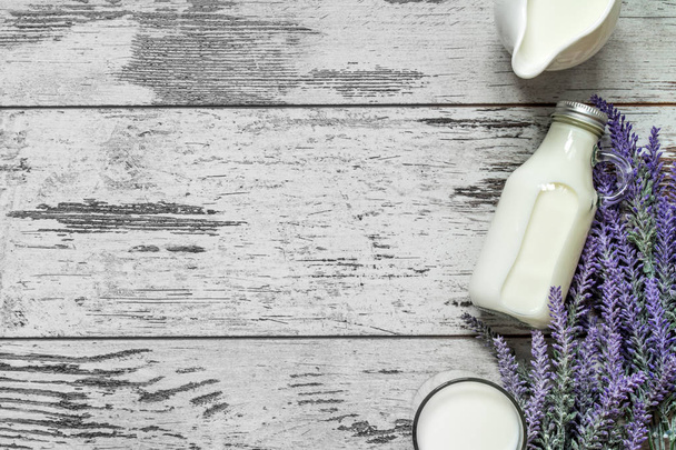 Vintage glass bottle with milk, a glass with milk and a beautiful white jug next to a branch of lavender flowers on a vintage wooden background. View from above. - Фото, изображение